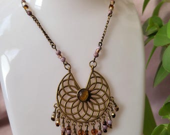 May include: A bronze-toned necklace with a filigree pendant and beaded accents. The pendant features a circular design with a central stone and dangling beads. The necklace is displayed on a white mannequin, with green foliage in the background.