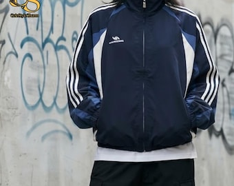 Puede incluir: Una chaqueta deportiva azul marino con cremallera, cuello alto, paneles de color blanco y azul claro en las mangas y tres rayas blancas a lo largo de cada brazo. La chaqueta incluye un pequeño logotipo blanco en el pecho.