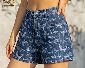 Puede incluir: Pantalones cortos de mezclilla de talle alto en un tono azul medio con un patrón de mariposas blancas dibujadas en líneas por toda la prenda. Los shorts incluyen un cierre de botón metálico plateado en la cintura, trabillas y cinco bolsillos.