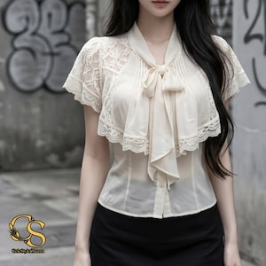 May include: A cream-coloured short-sleeve blouse featuring intricate lace detailing on the shoulders and sleeves, a front button closure, and a decorative neck tie with a ruffled collar. The garment includes the logo CeleStyleCharm in the corner.