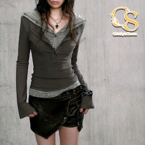 May include: An olive-green two-piece outfit. The top has long sleeves, a button-down front, and a layered design with a gray hood. The outfit includes a dark wrap-style skirt with a belt. The inner layer of the top has a pattern.