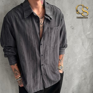 Puede incluir: Una camisa abotonada gris carbón con rayas verticales texturizadas. La camisa tiene un bolsillo en el pecho y un cuello clásico. Las mangas son largas y abotonadas en los puños. La camisa está desabrochada en la parte superior.