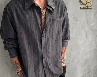 Puede incluir: Una camisa abotonada gris carbón con rayas verticales texturizadas. La camisa tiene un bolsillo en el pecho y un cuello clásico. Las mangas son largas y abotonadas en los puños. La camisa está desabrochada en la parte superior.