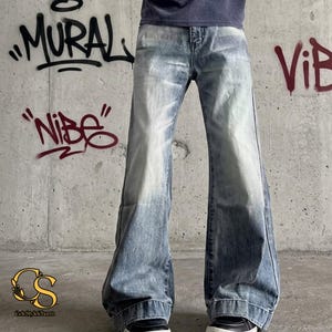 May include: Light blue denim jeans with a wide, flared leg. The jeans have a faded wash effect, with lighter areas on the thighs and knees. The background features graffiti art with the words "MURAL", "VIBE", and "Nibe".