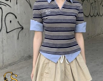 Puede incluir: Un polo de manga corta con rayas horizontales en azul marino, blanco y gris, con cuello y puños en azul claro. La prenda tiene un escote en V y el texto WITT HOY bordado en el pecho, combinado con una falda de color beige con cordón ajustable.