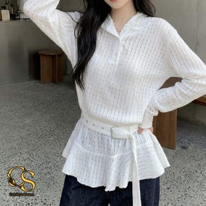 May include: A white long-sleeved shirt featuring a vertical ribbed texture, a collared neckline with a button placket, and a peplum-style hem. The garment includes a matching white fabric belt with a rectangular buckle cinched at the waist.