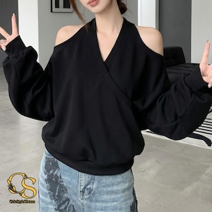 May include: A black long-sleeved sweatshirt featuring a V-neckline, a wrap-style front, and cold-shoulder cutouts. The garment has a relaxed fit with ribbed cuffs at the wrists and a ribbed hemline, paired with light blue denim jeans.