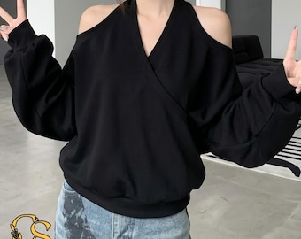 Puede incluir: Una sudadera negra de manga larga con escote en V, frente cruzado y aberturas en los hombros. La prenda tiene un corte holgado con puños y dobladillo acanalados, combinada con pantalones vaqueros de mezclilla en color azul claro.