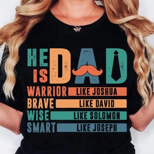 May include: Black t-shirt with the words "HE IS DAD" in teal, orange, and blue. The word "DAD" features an orange mustache and a teal tie. Below the text are the words "WARRIOR LIKE JOSHUA", "BRAVE LIKE DAVID", "WISE LIKE SOLOMON", and "SMART LIKE JOSEPH".