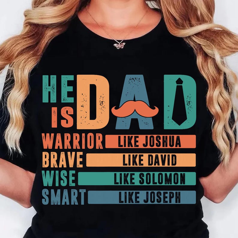 May include: Black t-shirt with the words "HE IS DAD" in teal, orange, and blue. The word "DAD" features an orange mustache and a teal tie. Below the text are the words "WARRIOR LIKE JOSHUA", "BRAVE LIKE DAVID", "WISE LIKE SOLOMON", and "SMART LIKE JOSEPH".