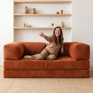 May include: A burnt orange corduroy sofa with a modern design. The sofa has a low profile and a comfortable appearance. The background features a white wall with wooden shelves displaying decorative pottery. A person is sitting on the sofa.
