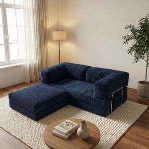 Puede incluir: Un sof&aacute; seccional de pana azul marino con una otomana, sobre una alfombra color crema. El sof&aacute; tiene un dise&ntilde;o moderno con un perfil bajo y una est&eacute;tica minimalista. Una peque&ntilde;a mesa de centro de madera con libros y un jarr&oacute;n est&aacute; frente al sof&aacute;.