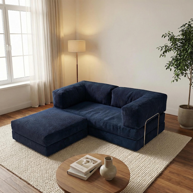 Puede incluir: Un sof&aacute; seccional de pana azul marino con una otomana, sobre una alfombra color crema. El sof&aacute; tiene un dise&ntilde;o moderno con un perfil bajo y una est&eacute;tica minimalista. Una peque&ntilde;a mesa de centro de madera con libros y un jarr&oacute;n est&aacute; frente al sof&aacute;.