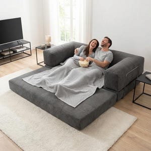 May include: A gray modular sofa with a textured surface, featuring a couple laughing and enjoying popcorn. A light gray blanket covers them. The room includes a black TV stand, side tables, and a white rug.