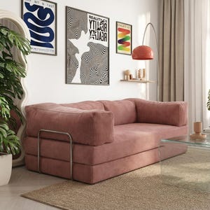 May include: A dusty rose corduroy sofa with a modern design. The sofa has a low profile, with a metal frame and a glass coffee table in front. The room features art prints and a floor lamp.
