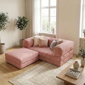 May include: A pink corduroy sectional sofa with an ottoman. The sofa is in a living room setting, with several throw pillows. The room includes a window, plants, and a neutral-toned rug. The sofa's dimensions are in centimeters.