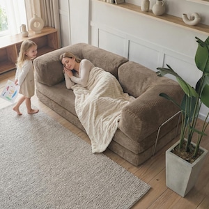 May include: A brown corduroy sofa with a woman sleeping under a cream-colored blanket. A young child walks nearby holding a drawing. A large potted plant sits to the right, and a neutral-toned rug covers the floor.