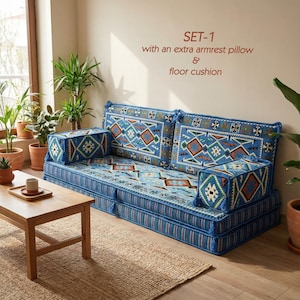 Puede incluir: Un juego de sofá de suelo azul con una almohada extra para el reposabrazos y un cojín de suelo. Los cojines presentan un estampado geométrico en tonos azules, naranjas y blancos. El sofá está colocado sobre una alfombra de yute, con una mesa de centro de madera y plantas en macetas cerca.