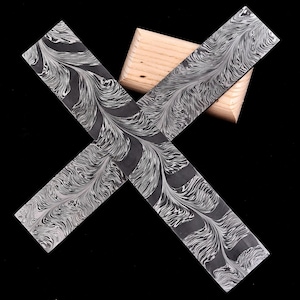 May include: Two rectangular pieces of steel with a swirling, feather-like pattern in shades of gray and white, resting on a small wooden block. The steel pieces are positioned in an X shape against a black background.