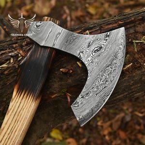 Puede incluir: Un hacha artesanal con una hoja de acero estampada y un mango de madera. La hoja tiene un diseño único y arremolinado. El mango es de madera clara con un acabado oscuro y quemado cerca de la cabeza. El hacha está sobre un trozo de madera.