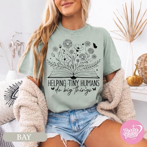 Puede incluir: Camiseta verde salvia con un diseño floral negro que emerge de un libro abierto, con el texto "Helping Tiny Humans do big things". La camiseta es de corte holgado y se combina con pantalones cortos vaqueros.