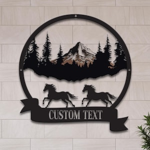 May include: Black metal wall art featuring a mountain scene with pine trees and two running horses inside a circular frame. A banner below reads "CUSTOM TEXT". The artwork is displayed on a light-colored tiled wall.
