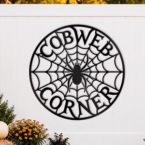 Op de afbeelding: Een rond wandbord van zwart metaal met een spinnenwebontwerp, een centrale spinnensilhouet en de tekst COBWEB CORNER geïntegreerd in het webpatroon. Het bord is bevestigd aan een witte schutting en heeft een uitgesneden ontwerp.