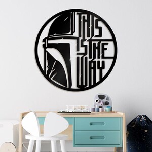 May include: Black metal wall art depicting a Mandalorian helmet inside a circle, with the words "THIS IS THE WAY" in bold letters. The artwork hangs on a white wall above a child's desk, with a light blue and wooden desk.