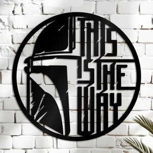 May include: Circular black metal wall art with a helmet design and the words "THIS IS THE WAY". The artwork is mounted on a white brick wall. The design is a bold, stylized statement piece.