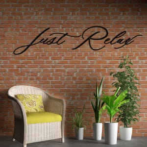 May include: A black metal sign that reads "Just Relax" is mounted on a red brick wall. A white wicker chair with a yellow cushion and floral pillow sits in front of the wall. Potted plants in white pots are arranged to the right of the chair.