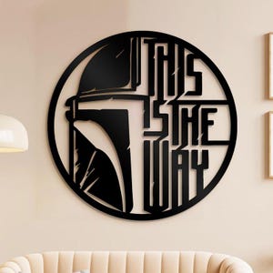May include: Black metal wall art with a circular design. It features a helmet silhouette and the text "THIS IS THE WAY." The artwork is mounted on a light-coloured wall, creating a striking contrast. The design is a popular theme.