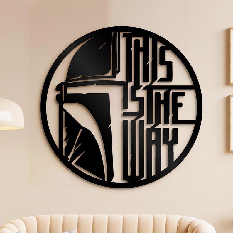 May include: Black metal wall art with a circular design. It features a helmet silhouette and the text "THIS IS THE WAY." The artwork is mounted on a light-coloured wall, creating a striking contrast. The design is a popular theme.