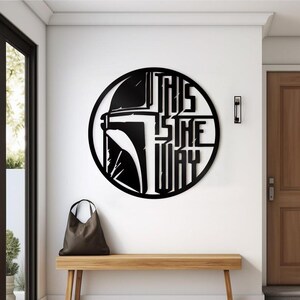 May include: Black metal wall art depicting a Mandalorian helmet and the words "THIS IS THE WAY" in a circular design. The artwork is mounted on a white wall above a wooden bench. A brown tote bag rests on the bench.