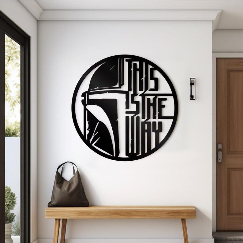 May include: Black metal wall art depicting a Mandalorian helmet and the words "THIS IS THE WAY" in a circular design. The artwork is mounted on a white wall above a wooden bench. A brown tote bag rests on the bench.