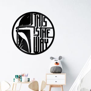 May include: Circular black metal wall art displaying a Mandalorian helmet and the words "THIS IS THE WAY". The design is mounted on a white wall. The artwork is suitable for a child's room.
