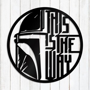 May include: Black metal wall art with a helmet design and the words "THIS IS THE WAY" in a circular frame. The artwork is set against a white wooden background.