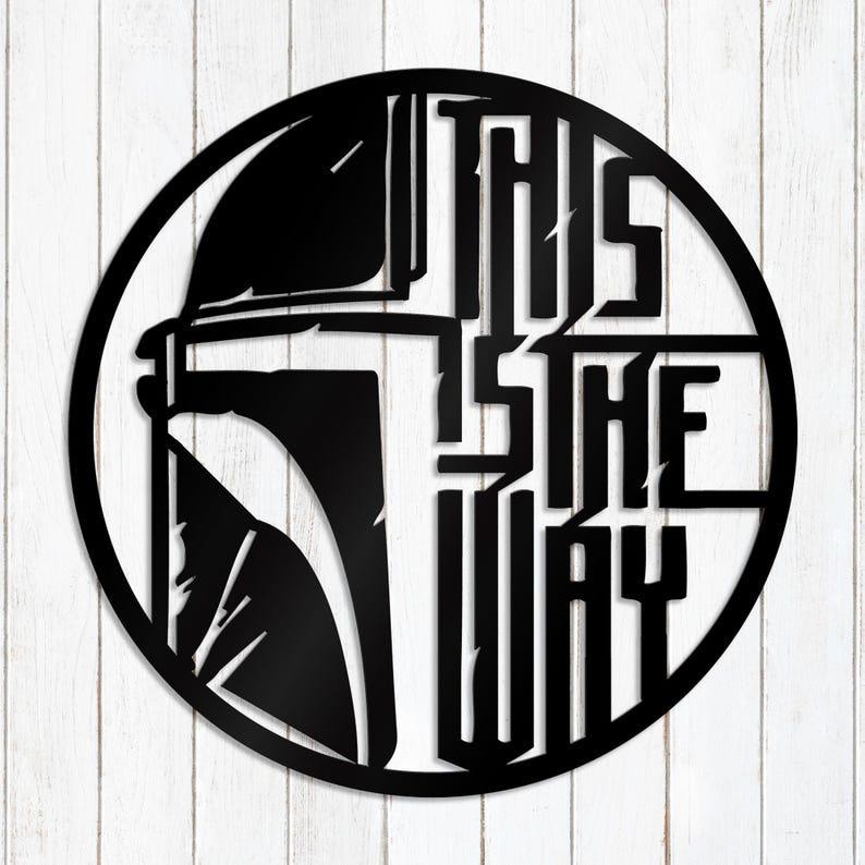 May include: Black metal wall art with a helmet design and the words "THIS IS THE WAY" in a circular frame. The artwork is set against a white wooden background.