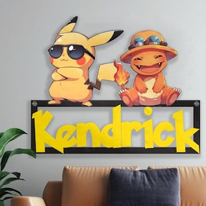 May include: A black framed wall decoration featuring yellow Pikachu and Charmander characters. The name "Kendrick" is displayed in yellow letters below the characters. The Pikachu character is wearing sunglasses.