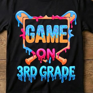 May include: A black short-sleeve t-shirt featuring a graphic design with the text "GAME ON 3RD GRADE" in a dripping, colorful font. The design includes a baseball home plate shape and two crossed baseball bats in orange and blue.