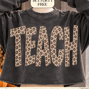 May include: A charcoal grey cropped sweatshirt featuring the word TEACH in large, bold letters with a tan, brown, and black leopard print pattern. The garment has long sleeves and a raw, unfinished hemline. A small label at the top reads BUY 1 GET 1 FREE.