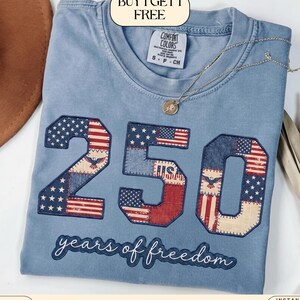 Puede incluir: Una camiseta Comfort Colors azul claro con un diseño patriótico. El número "250" se muestra, incorporando patrones de la bandera estadounidense. El texto "years of freedom" está debajo. Se ve un collar dorado.