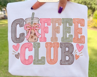 May include: A white t-shirt featuring a graphic design with the text COFFEE CLUB in multi-coloured, layered line art. The letter O is replaced by an iced coffee cup with a pink bow. Small pink and brown hearts surround the text.