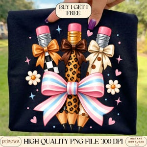 May include: A black t-shirt featuring a graphic of three pencils with pink erasers, each adorned with a small bow. The pencils have checkered, leopard, and polka dot patterns, tied together by a large pink, white, and blue bow. Text reads BUY 1 GET 1 FREE.