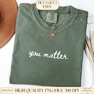 Puede incluir: Una camiseta verde oliva con las palabras "you matter" en escritura blanca. Un collar plateado con un pequeño colgante descansa sobre la camiseta. La camiseta tiene una etiqueta Comfort Colors y está doblada. Un sombrero marrón es parcialmente visible.