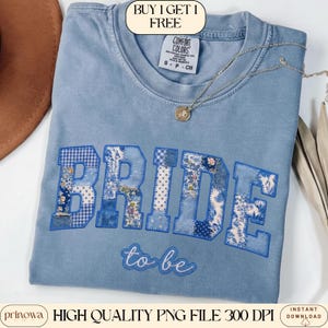 Puede incluir: Una camiseta Comfort Colors azul claro con la palabra "BRIDE" en letras de patchwork. Las letras son de varios tonos de azul y patrones florales. Debajo de la palabra "BRIDE" está la frase "to be". Un collar plateado con un colgante está en la camiseta.
