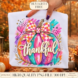 May include: A white t-shirt featuring a colorful graphic of a pumpkin with a checkered orange and white bow. The pumpkin is painted in vibrant teal, pink, and orange brushstrokes with gold glitter accents and the word thankful in white script.