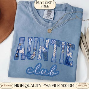 May include: A light blue Comfort Colors t-shirt with the words "AUNTIE Club" in a patchwork design. The word "AUNTIE" is in a collegiate style font with floral and gingham patterns. A silver necklace with a pendant is draped on the shirt.