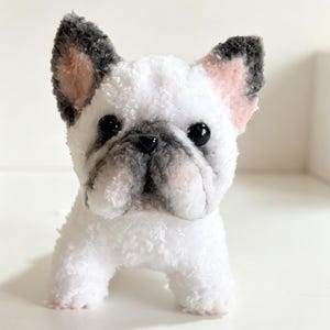 以下が含まれることがあります： 耳と口元に黒とグレーのアクセントが付いた、白いフレンチブルドッグのぬいぐるみ。おもちゃは黒い目とピンクの内耳を持っています。犬は四本足で立っています。