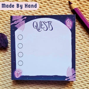 May include: A square notepad with a dark purple starry border and the word Quests printed at the top. The centre is a light purple arch shape with four empty circular checkboxes. The design features illustrations of light purple dragon wings and crystals.