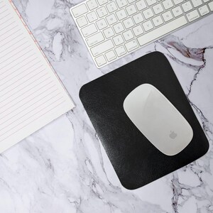 Può includere: Un mouse wireless bianco con il logo Apple è appoggiato su un tappetino nero. Sono visibili anche una tastiera bianca e un blocco note con linee rosa. Gli oggetti sono disposti su una superficie con motivo marmoreo.